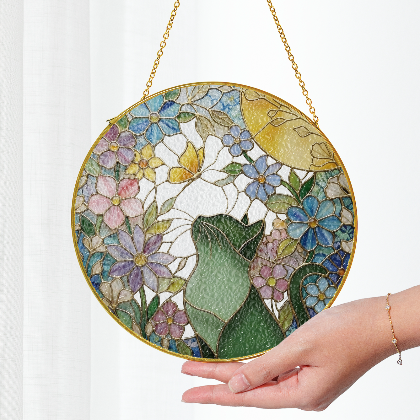 Round gold stained glass sun catcher with of a cat looking at a butterfly floral design held by a hand on a white background