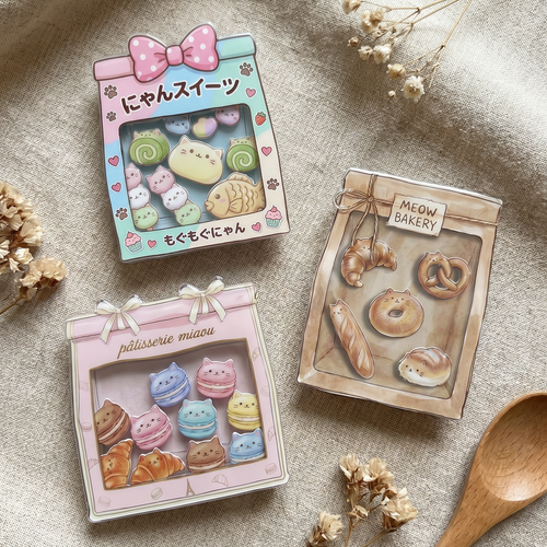 Three shaker phone grips of bakery bags with cute cat designs on a textured surface with dried flowers and a wooden spoon.