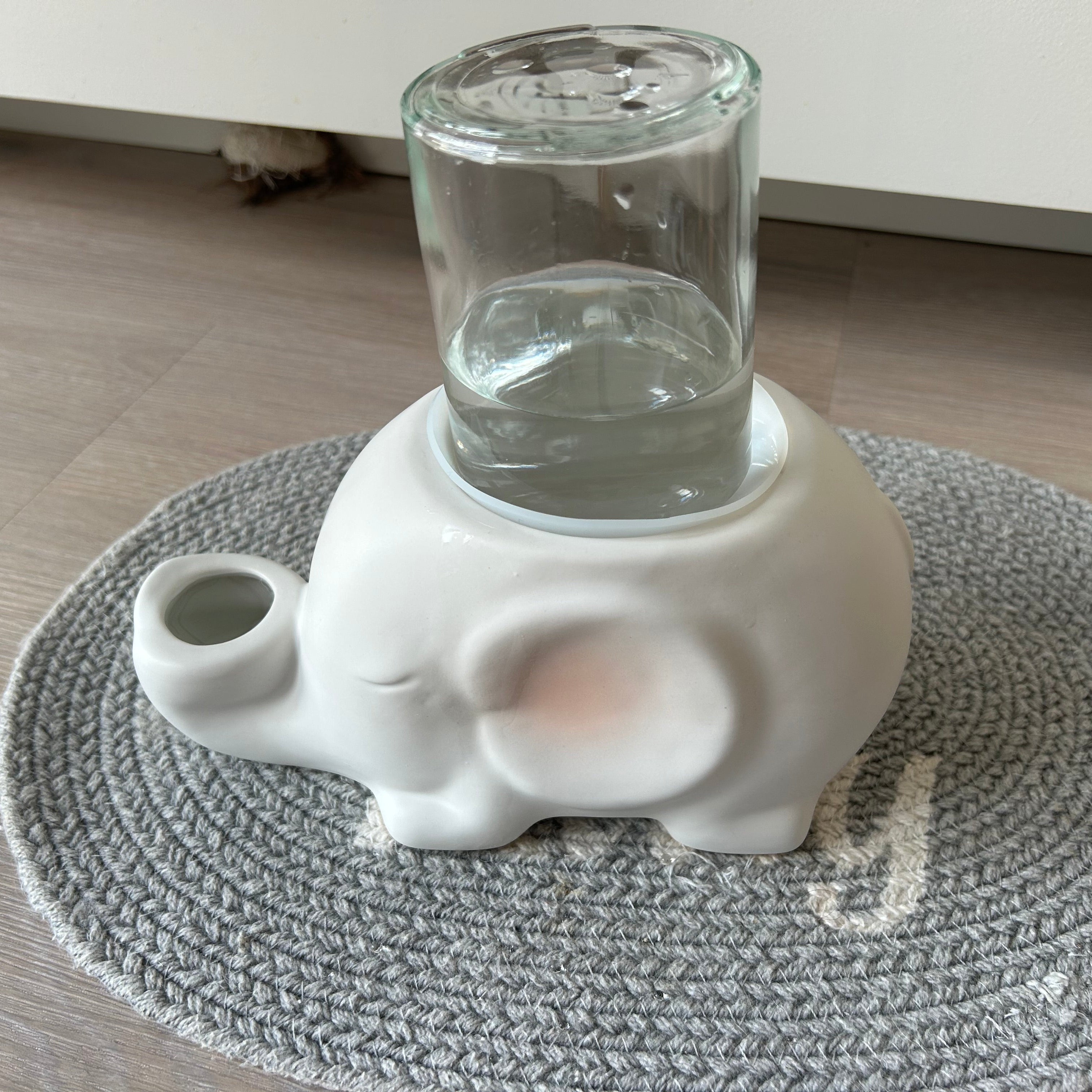 White elephant-shaped gravity water bowl for no wet cat chins with a glass containing water on a gray textured mat.