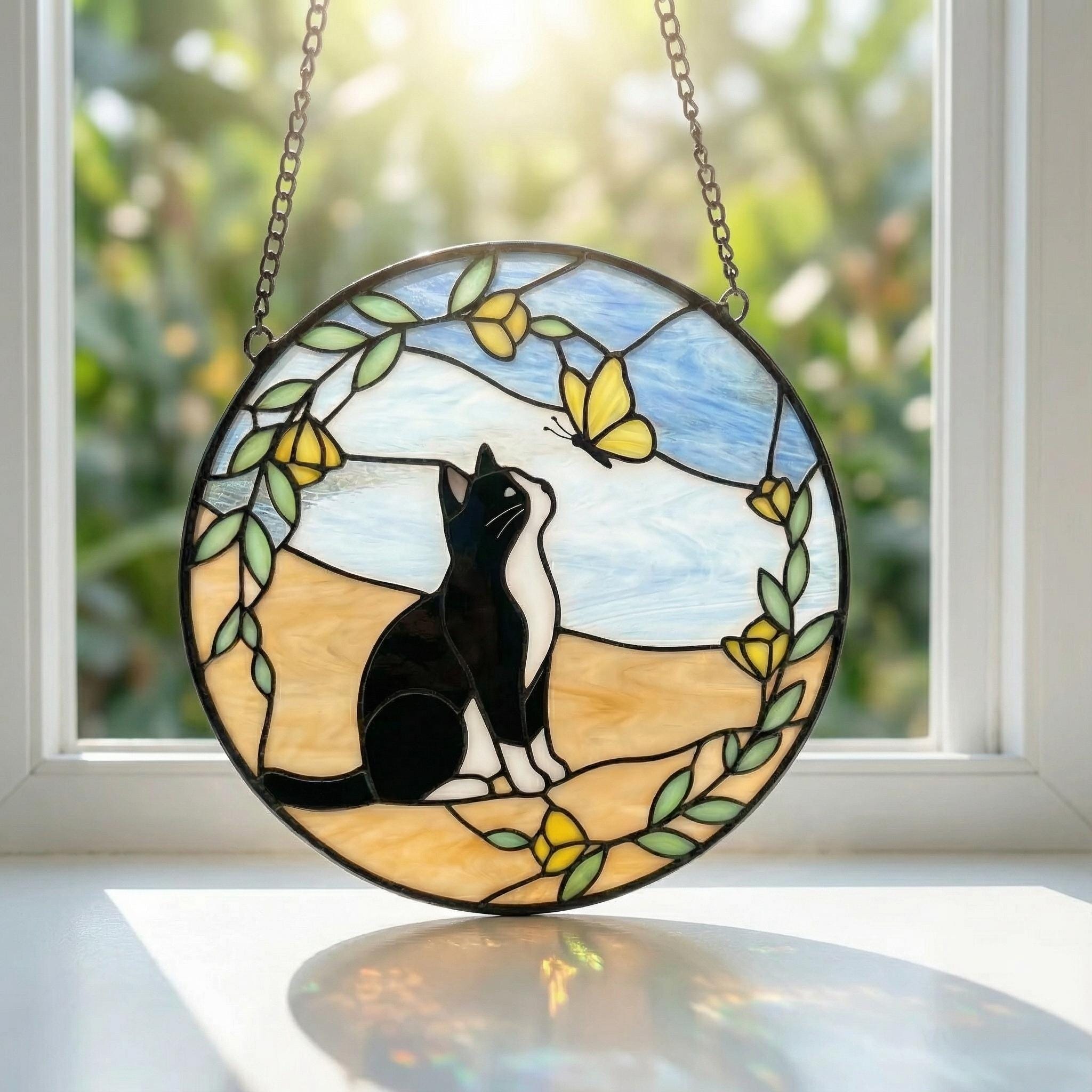 Stained glass sun catcher  with a tuxedo cat and butterfly design on a windowsill.