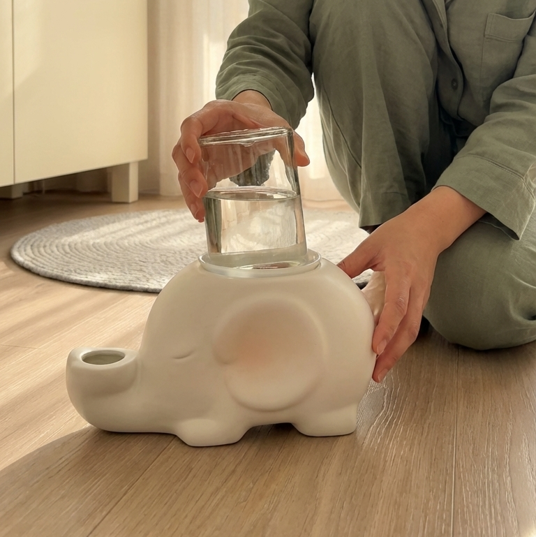Woman kneeling on the floor with a white elephant-shaped gravity glass and ceramic water bowl for no wet cat chins.